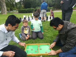 Two dads play a board game in the park as their children watch on. All socially distanced.