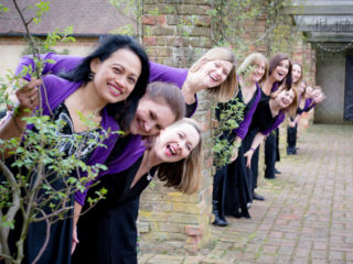 12 members of the Military Wives Choirs pose for a photo wearing choir outfits including a black dress and purple cardigan