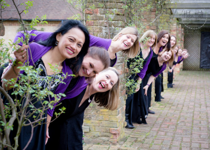 12 members of the Military Wives Choirs pose for a photo wearing choir outfits including a black dress and purple cardigan