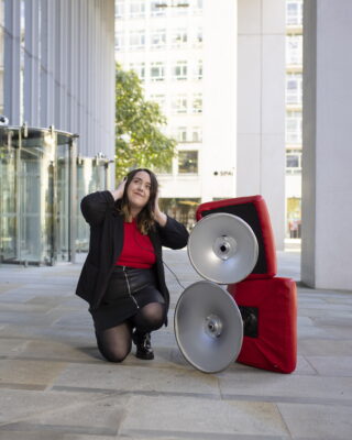 A picture of Ashley Holding posing next to a couple steel pan speakers, whilst holding her headphones, listening to music