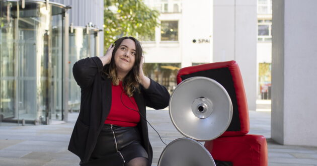 A picture of Ashley Holding posing next to a couple steel pan speakers, whilst holding her headphones, listening to music